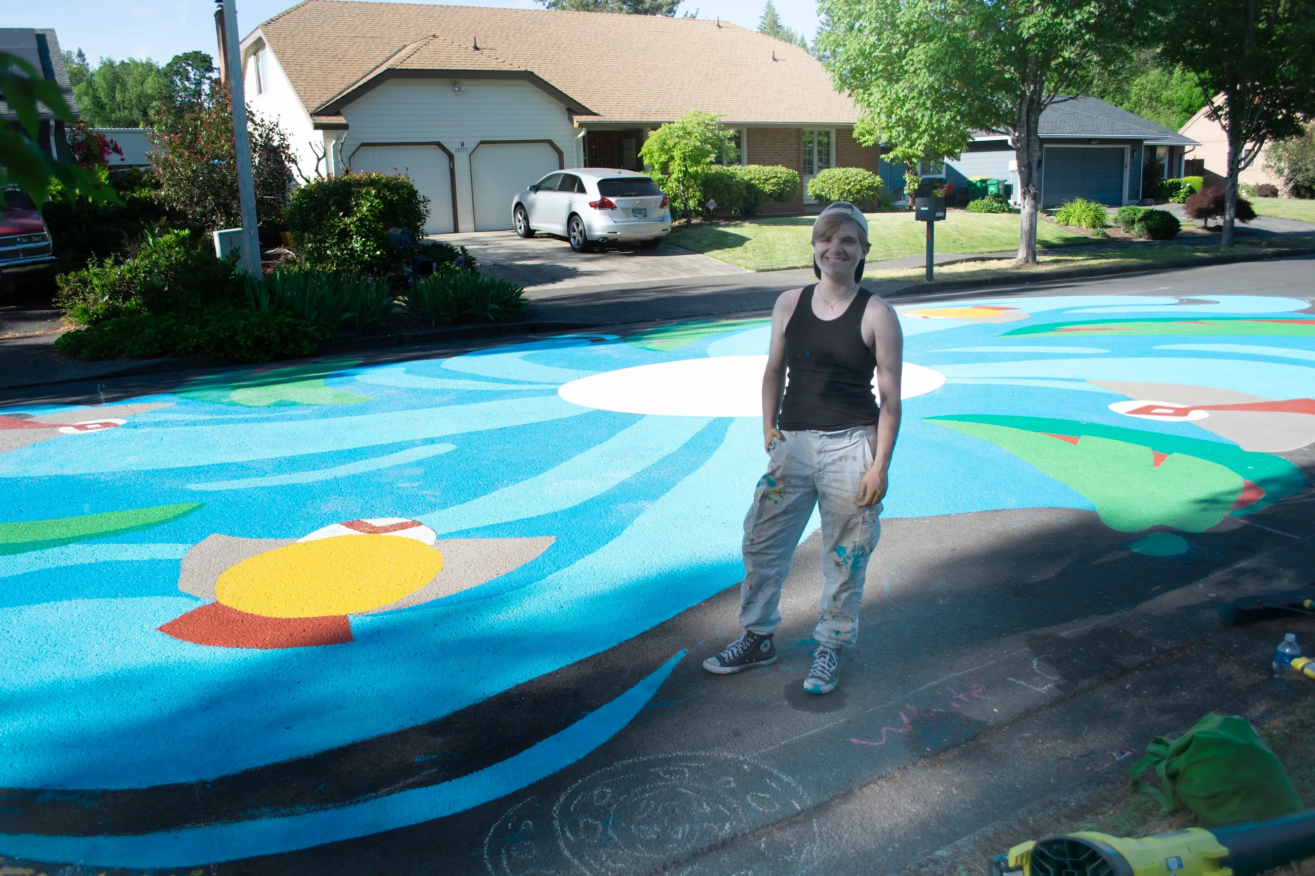 Everett Liddle standing on the completed street mural, a swirl of blue water, green leaves, and wildlife painted across a full neighborhood intersection.