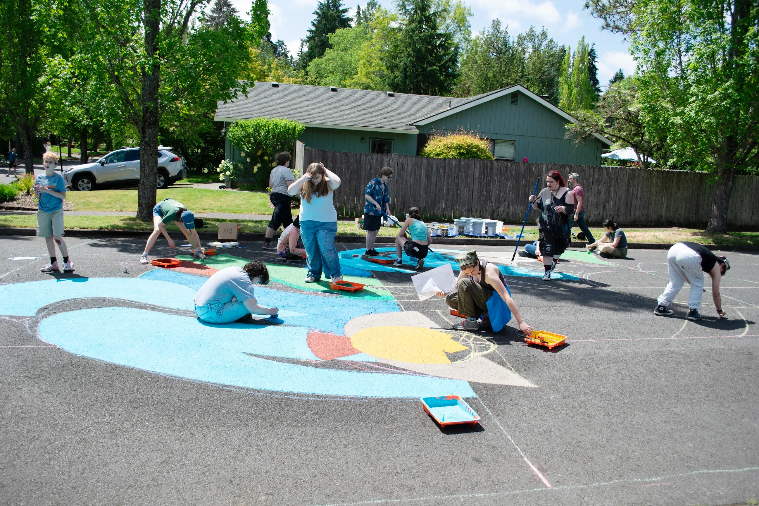 Student volunteers spread across the intersection painting the mural, filling in the blue swirl and green leaves with rollers and trays.
