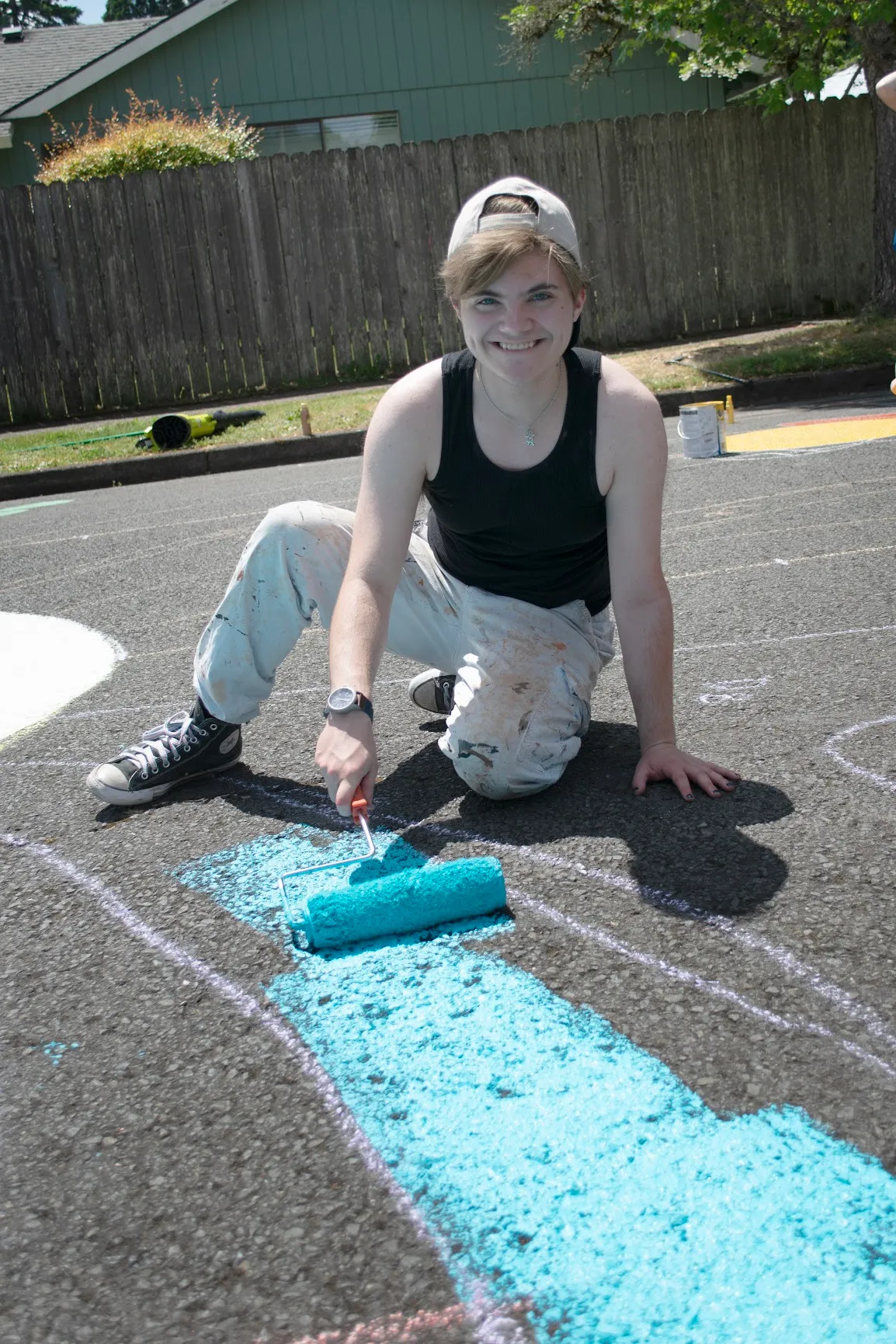 Everett kneeling on the closed-off street mid-installation in a black tank top, paint-spattered light jeans and a backwards cap, smiling at the camera as he runs a roller loaded with turquoise across the asphalt, with freshly painted turquoise bands and white chalk outlines around him and a wood fence and green house behind.